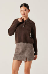 A woman is wearing a dark brown, chunky knit sweater with a cable pattern. The sweater features a collared neckline with four decorative buttons, each adorned with a flower-like design, and long sleeves. She also wears a beige and brown herringbone patterned skirt with a small slit in the front.